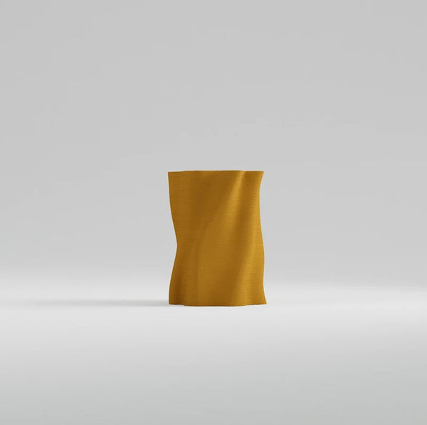 A modern, sculptural vase made of wavy, draped fabric in a rich mustard yellow color, set against a seamless light gray studio background.